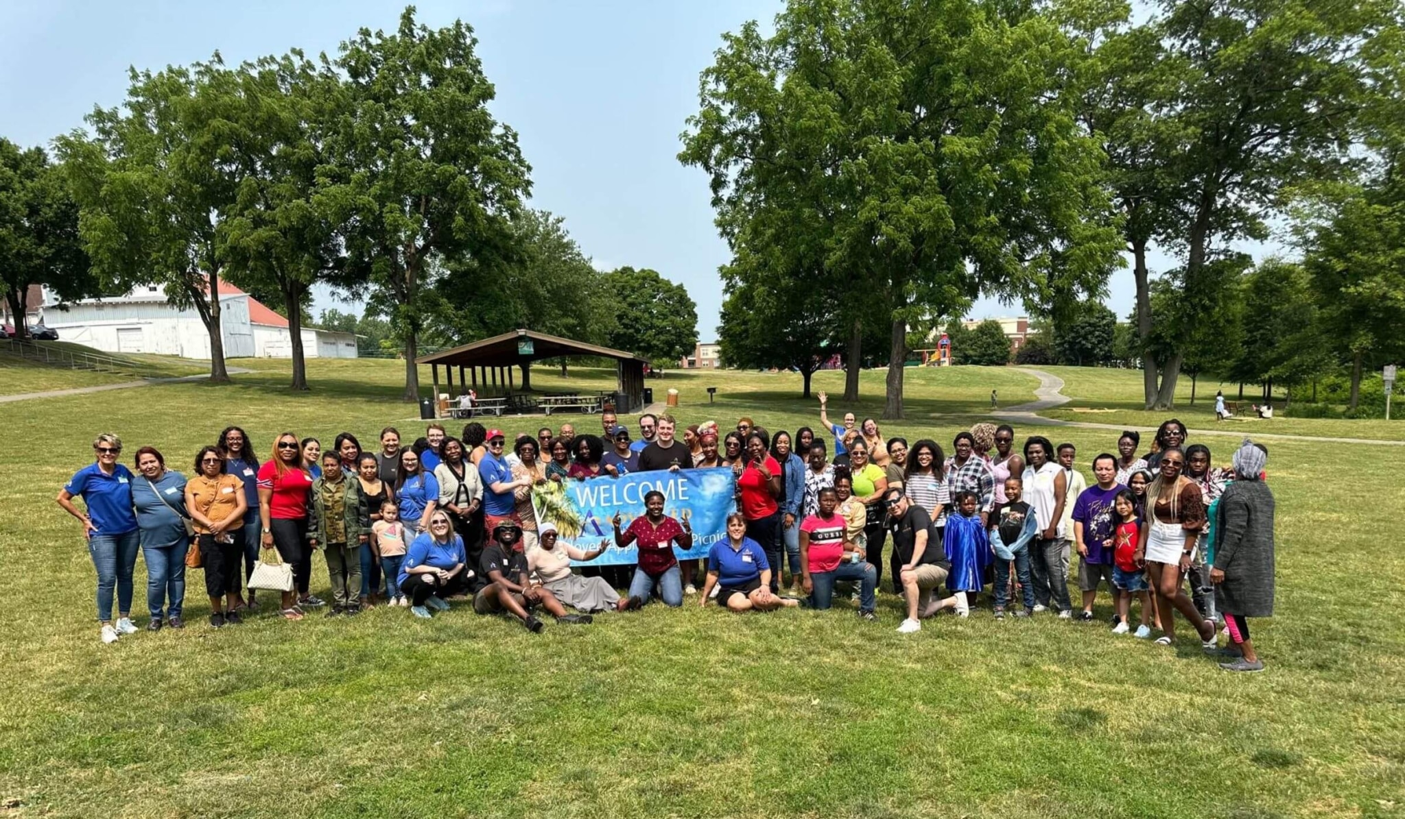 Large group photo of Advanced Nursing & Home Care team and families at a summer picnic in a park, celebrating unity and care.