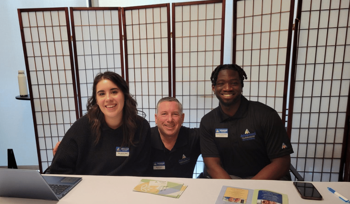 Three smiling team members from Advanced Nursing & Home Care sitting together at a community event table, representing trusted senior care services.