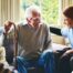 A caregiver is smiling and supporting an elderly man with a cane while another senior woman looks on, showing trust and compassionate home care.
