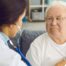 A nurse uses a stethoscope to check the heart of an elderly man at home, representing the professional medical support offered by Advanced Nursing & Home Care.