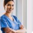 A smiling caregiver in blue scrubs stands confidently with arms crossed, representing the compassionate and professional care at Advanced Nursing & Home Care.